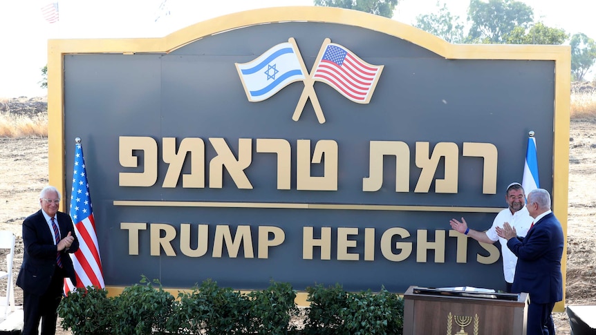 A man in a suit applauds near a large sign with the words "Trump Heights" written under Hebrew text.