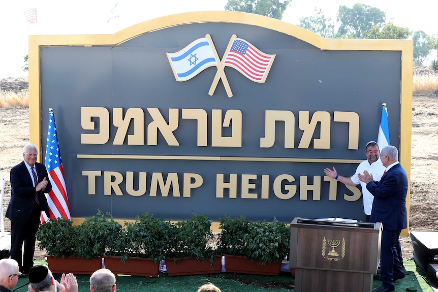 A man in a suit applauds near a large sign with the words "Trump Heights" written under Hebrew text.