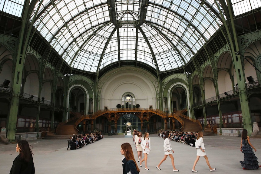 Models walk through an enormous historic room with a glass roof and green metal pillars