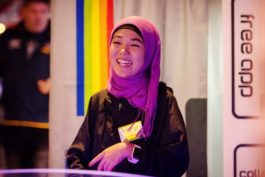 Rahila smiles as she looks at someone opposite her. She wears a purple head scarf and stands in front of a rainbow curtain.