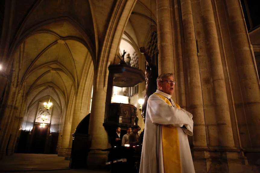 Patrick Chauvet looks straight ahead as he walks through a dimly lit cathedral. He wears white robes with a yellow scarf.