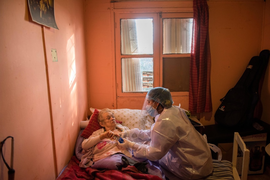 A woman dressed in personal protective equipment including a mask and gloves tends to a woman lying on a bed.