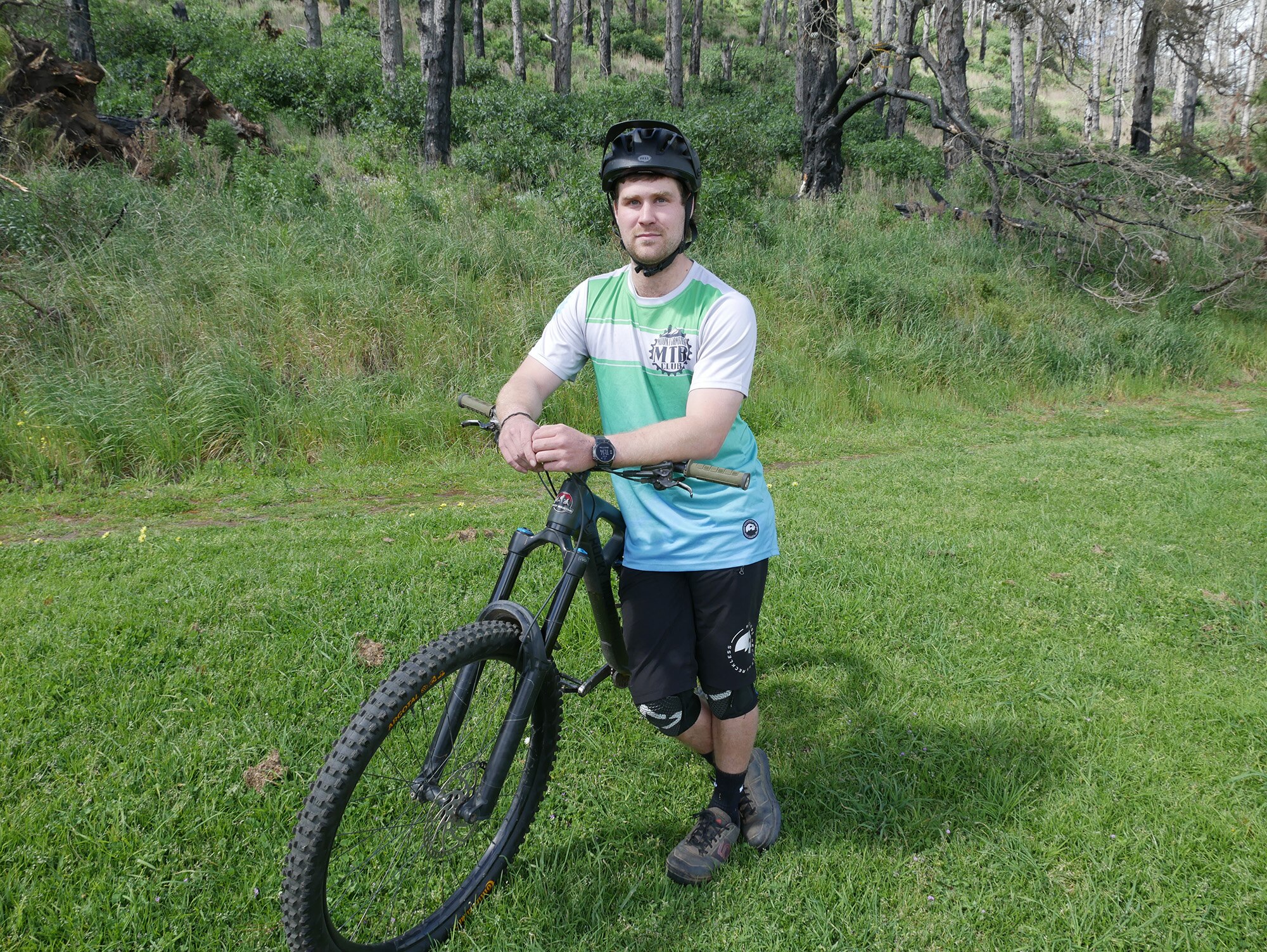 A white man with a helmet on stands with a yellow mountain bike on a nature path in front of a fallen, burnt pine tree.