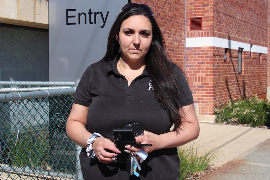 A mid shot of a middle aged woman with dark hair posing for a photo outside a school holding keys and a phone.
