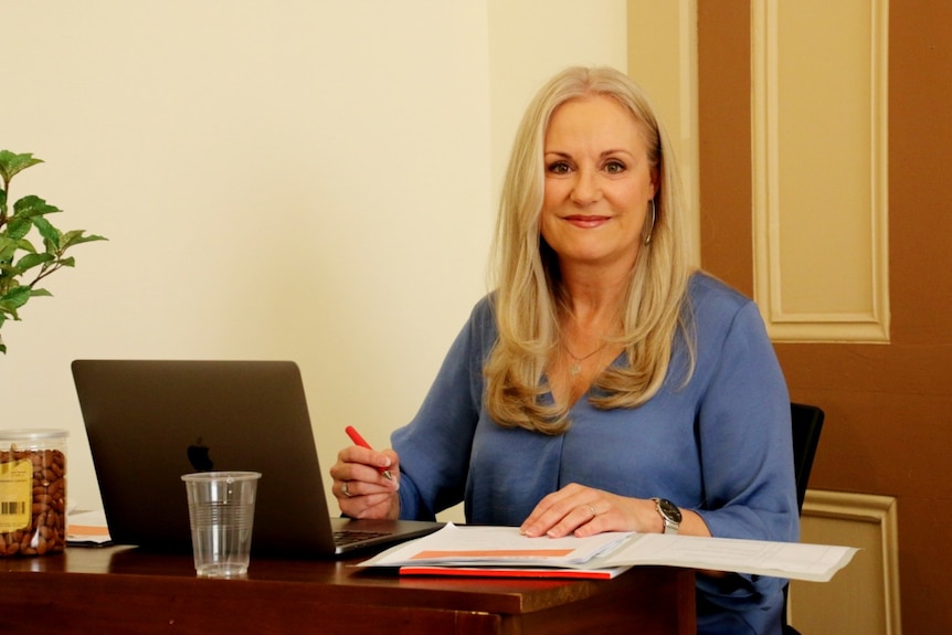 A woman with blonde hair wearing a blue top sits at a desk holding a pen in front of a laptop.