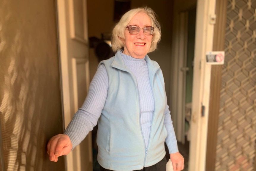 A light-haired woman wearing glasses stands on her doorstep and looks into the camera.