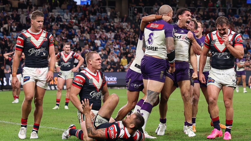 Melbourne Storm players jump, shout and celebrate a try as Roosters defenders look dejected. 
