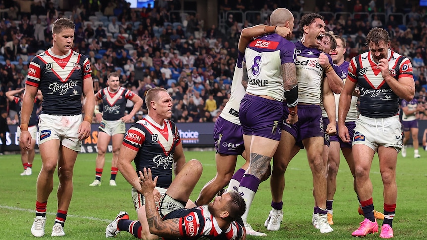 Melbourne Storm players jump, shout and celebrate a try as Roosters defenders look dejected. 