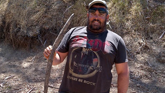A man with dark facial hair, a cap, and an AC/DC shirt stands in a dry creek bed, holds a curved piece of wood about 2 foot long