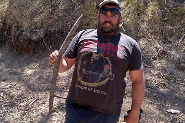 A man with dark facial hair, a cap, and an AC/DC shirt stands in a dry creek bed, holds a curved piece of wood about 2 foot long