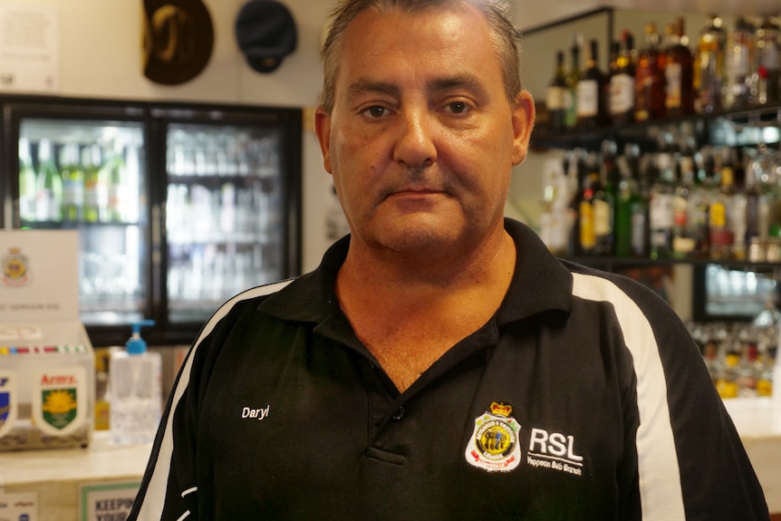 An older man in a polo shirt looks at the camera with a straight face, bar fridges and drinks behind.