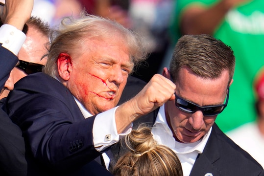 Donald Trump, bleeding from the ear, raises a fist while surrounded by bodyguards.