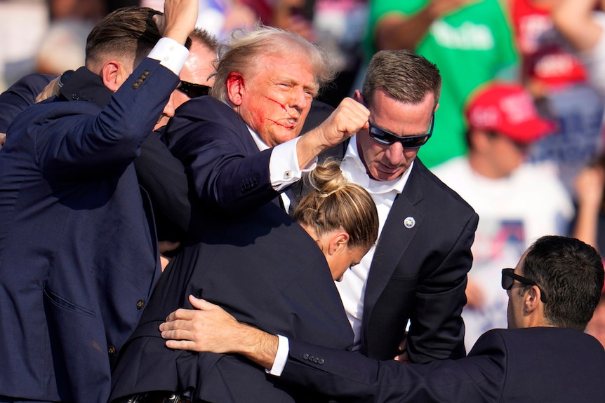 Donald Trump, bleeding from the ear, raises a fist while surrounded by bodyguards.