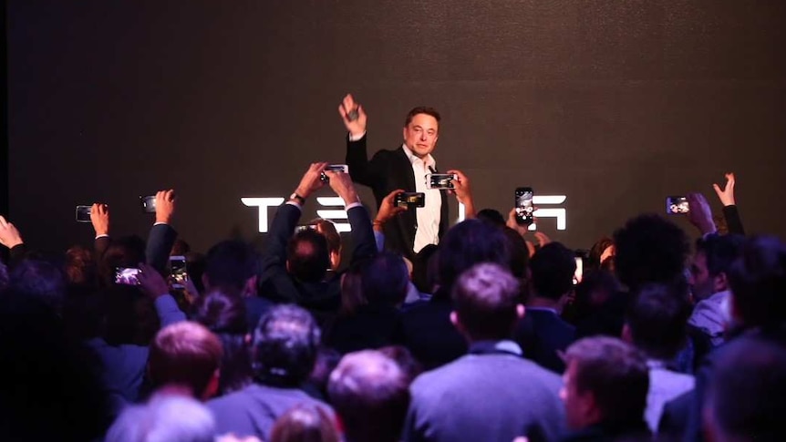 Elon Musk stands in front of a giant screen, talking to the audience