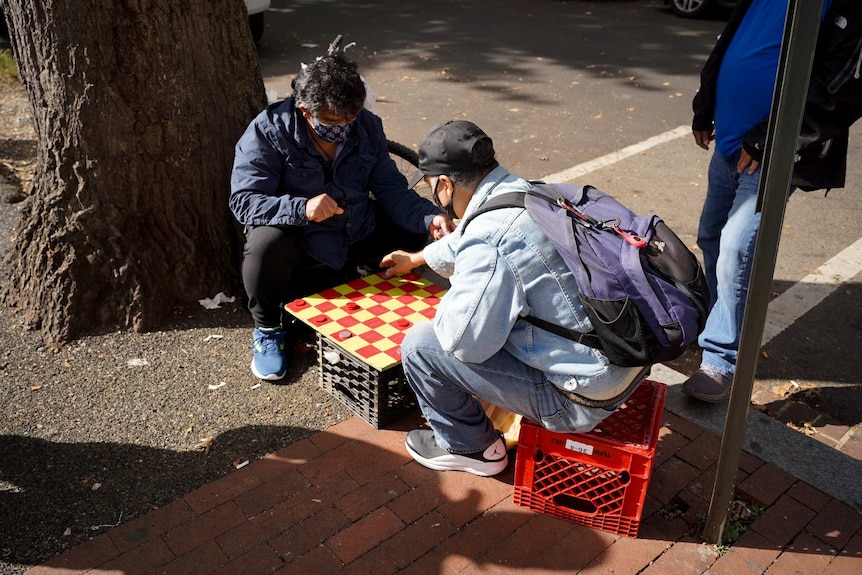 Two men wearing masks and jackets sit on milk crates hunched over a red and yellow checker board as another man watches on