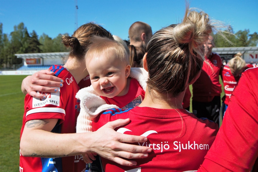 Women in red football jerseys stand in a  circle, their backs turned. Close up of toddler girl's smiling face 