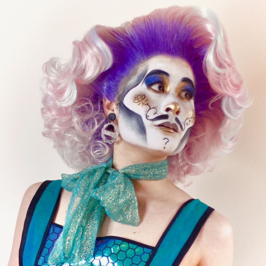 Randy Roy in a pink and purple wig, elaborate makeup and a blue scarf.
