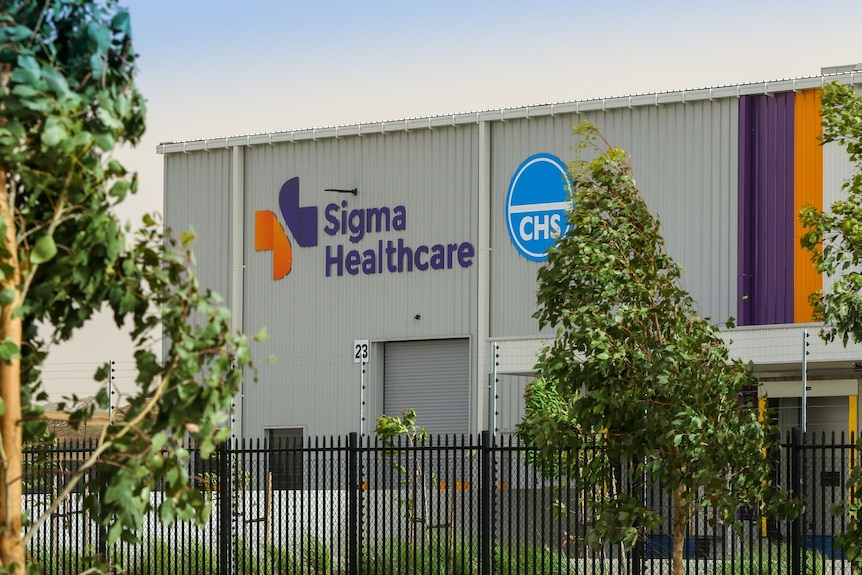 A large beige warehouse behind a black fence with Sigma Healthcare written on the side of the building in purple letters.