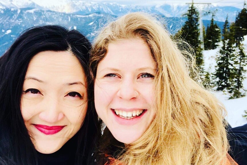 An Asian woman and a western woman hug and smile to the camera in snowy mountains.