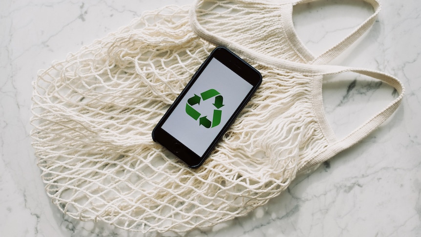 a white cotton mesh bag with a phone on top showing the green recycle symbol