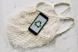 a white cotton mesh bag with a phone on top showing the green recycle symbol