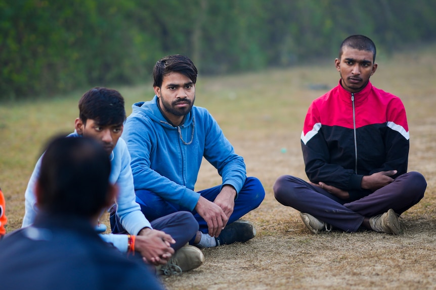 Men sit cross-legged next to each other in a circle, one in blue hooded jumper and another in red and black