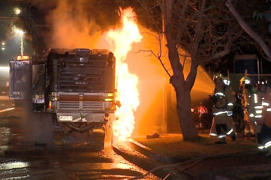 A road milling machine is on fire, a firefighting crew hose the vehicle down.