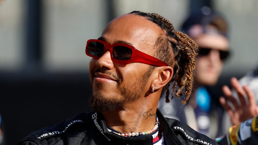 Lewis Hamilton, in his race suit, no helmet, wearing sunglasses, looking upward with a smile in the paddock