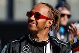 Lewis Hamilton, in his race suit, no helmet, wearing sunglasses, looking upward with a smile in the paddock