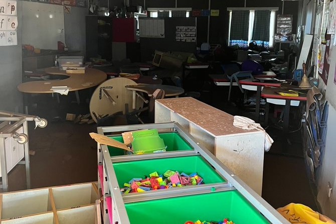 A cluttered room with tables overturned, shelving with drawers containing lego on its side.