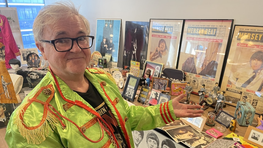 A man in a lime green suit jacket with red details gestures to a large table of Beatles merchandise.