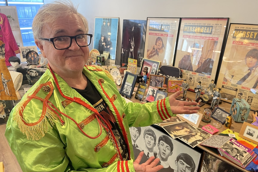 A man in a lime green suit jacket with red details gestures to a large table of Beatles merchandise.