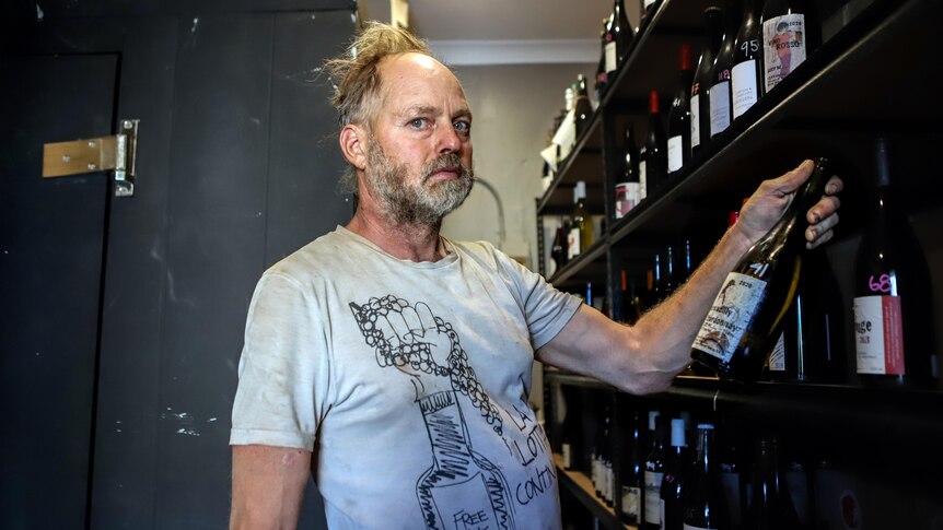 Man with thinning straw blonde hair and grey t-shirt holds bottle of wine inside wine cellar with shelf of wines to his side
