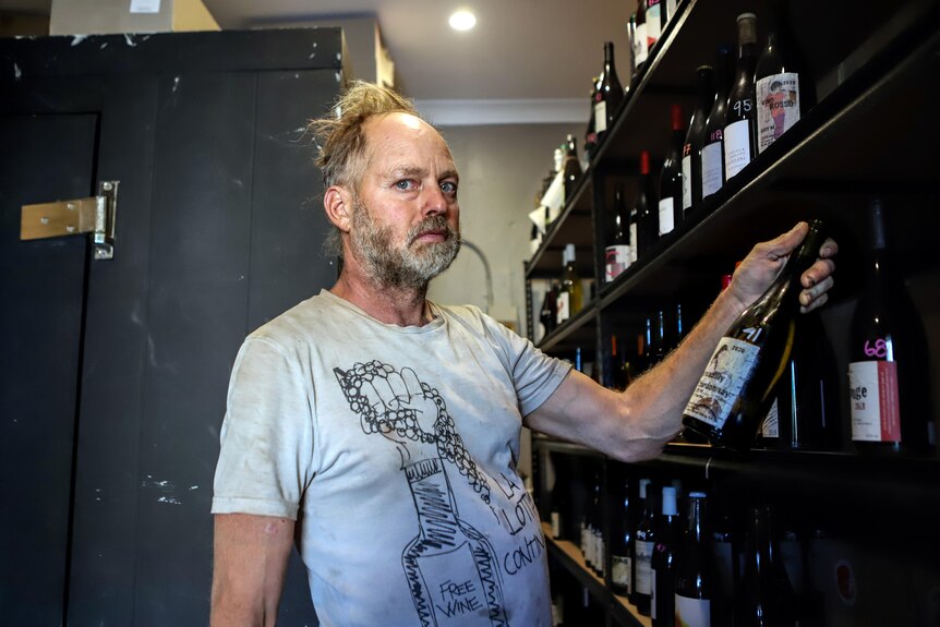 Man with thinning straw blonde hair and grey t-shirt holds bottle of wine inside wine cellar with shelf of wines to his side