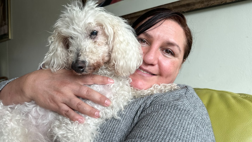 A woman with brown hair and brown eyes, cuddles a white curly haired poodle, sitting on a green couch. 