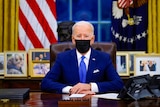 Joe Biden in a suit and black face mask sitting at the Resolute Desk in the Oval Office