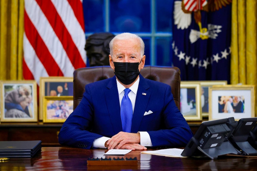 Joe Biden in a suit and black face mask sitting at the Resolute Desk in the Oval Office
