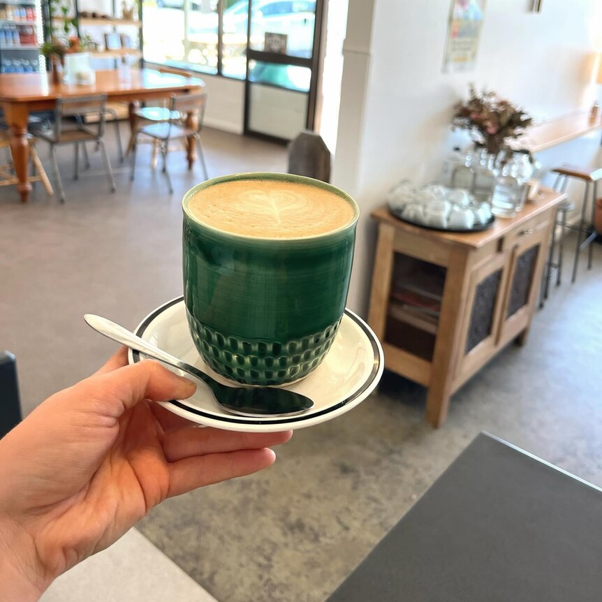 A hand holds out a coffee in a handmade ceramic cup, cafe in background