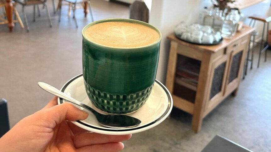 A hand holds out a coffee in a handmade ceramic cup, cafe in background