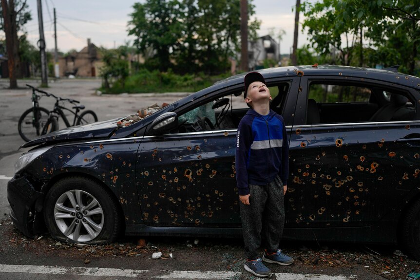 Little boy stands in front of a bullet marked navy blue car looking up