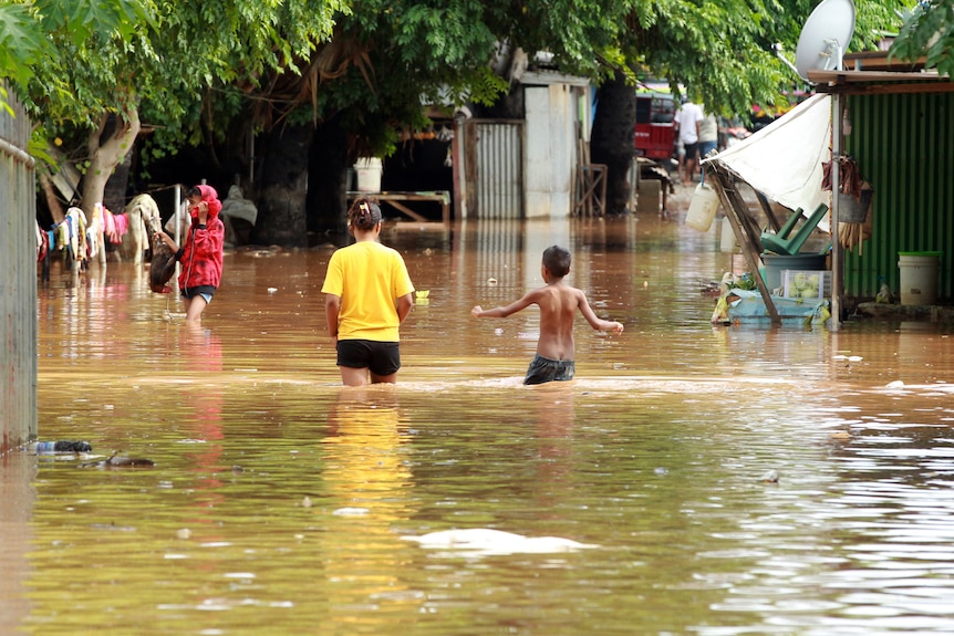 A woman and a boy walk through brown, waist-high floodwaters along what is usually a street.