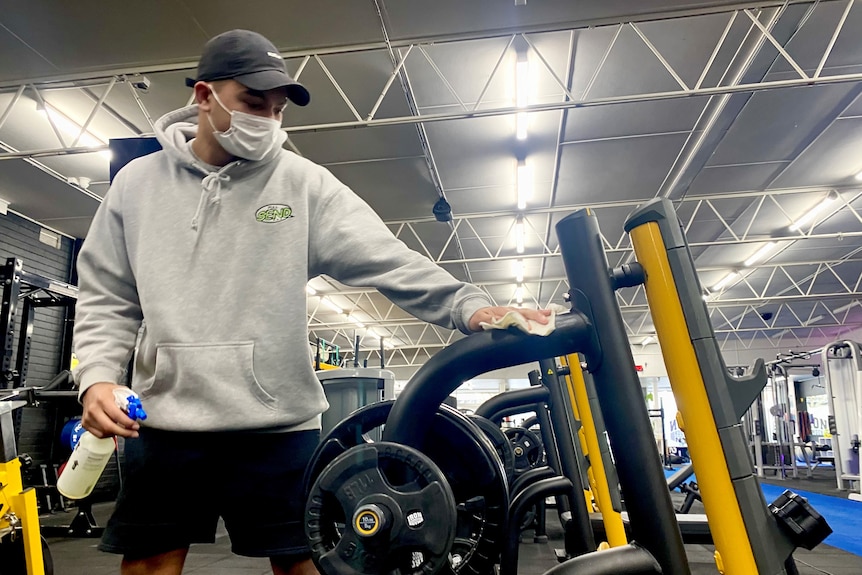 Un homme portant un sweat à capuche gris et un masque désinfecte l'équipement de gym.