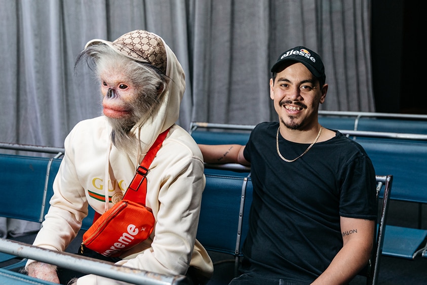 A smiling man with cap, black tee and gold chain sits on airport style seats next to Yunnan snub nosed monkey in human clothes.