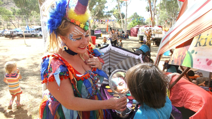 woman dressed up in colour costume holds out hand mirror for child with face painted.