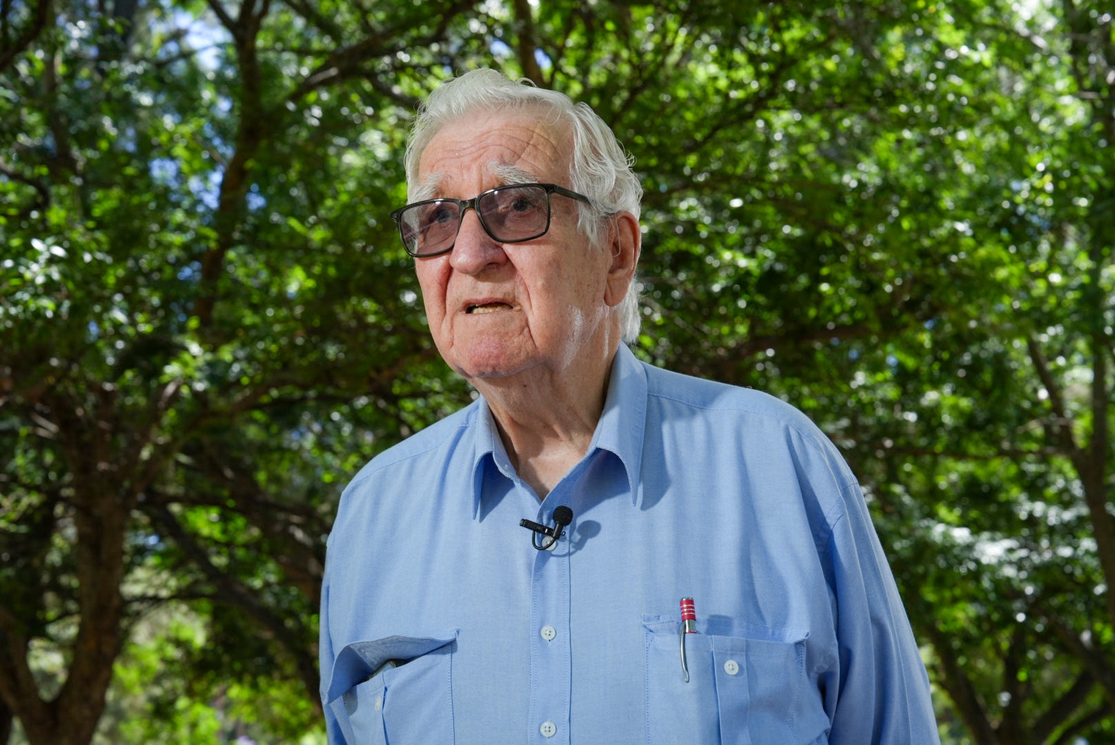An older man wearing sunglasses and a longsleeve blue shirt with a pen in his pocket stands in front of trees.