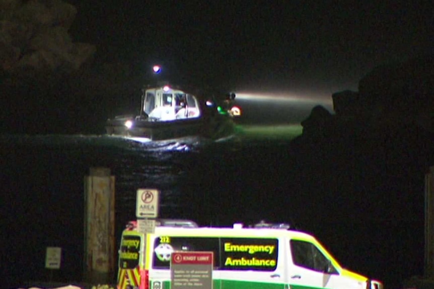 A boat with a spotlight leaves from a dock with an ambulance on it