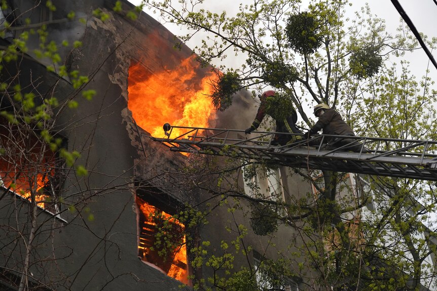 Firefighters on a ladder work to extinguish fire coming out of a window at an apartments building. 