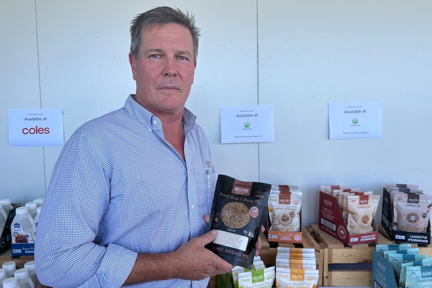 A middle aged man is holding a packet of seeds and grain mix standing in front of a table filled with branded products