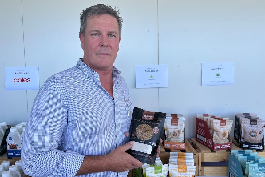 A middle aged man is holding a packet of seeds and grain mix standing in front of a table filled with branded products