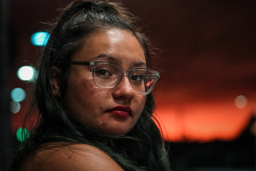 A close-up of Tee looking into the camera, with a dark red sunset behind her.